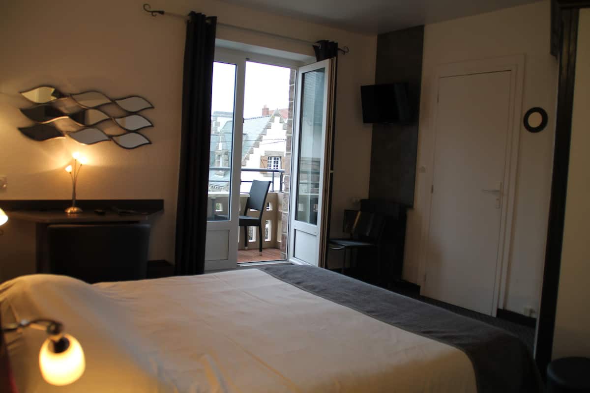A hotel room with a large bed, a desk with a lamp, a chair, a TV mounted on the wall, and a balcony with a chair and table.
