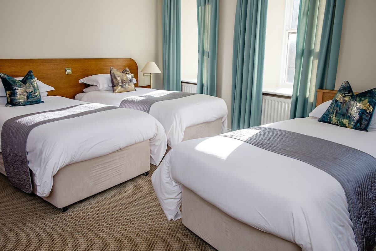 A hotel room with three beds, a wooden headboard, a lamp, and light blue curtains.