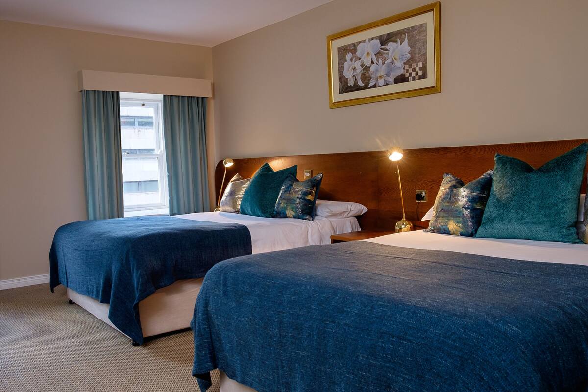 A hotel room with two beds, each with a blue bedspread and matching pillows, a wooden headboard, a window with curtains, and a framed picture on the wall.