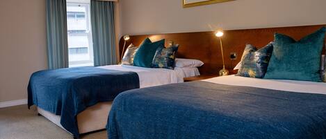 Habitación de hotel con dos camas, cada una con cubrecama y almohadas azules a juego, cabecera de madera, ventana con cortinas e imagen enmarcada en la pared.