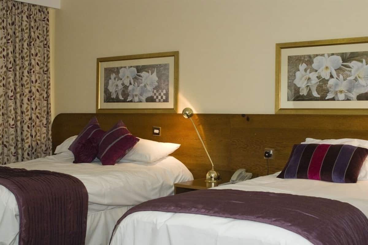 A hotel room with two beds, a wooden headboard, a bedside lamp, and two framed floral paintings on the wall.