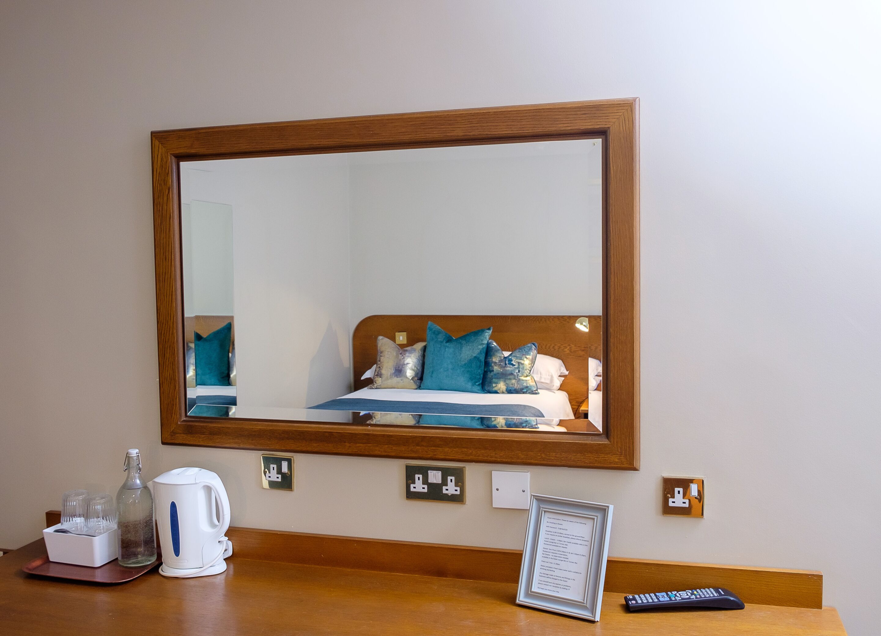 Quarto de hotel com um espelho grande, cama com roupa de cama azul e branca, mesa de cabeceira com chaleira, copos e controle remoto.