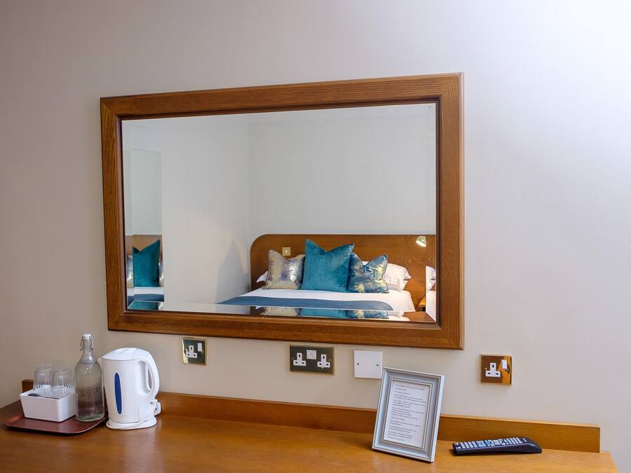 Quarto de hotel com um espelho grande, cama com roupa de cama azul e branca, mesa de cabeceira com chaleira, copos e controle remoto.