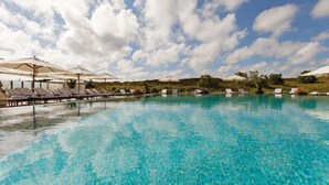 A clear blue pool with lounge chairs and umbrellas, surrounded by greenery under a partly cloudy sky.