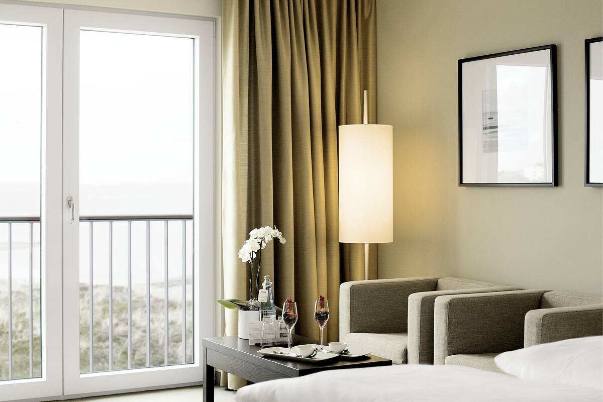 A hotel room with a bed, a seating area with two sofas, a table with a wine glass and flowers, a lamp, and a view of the outdoors through a sliding door.