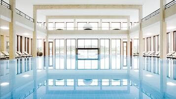 Una amplia piscina interior con agua cristalina y azul, rodeada de tumbonas y grandes ventanales con vistas al exterior.