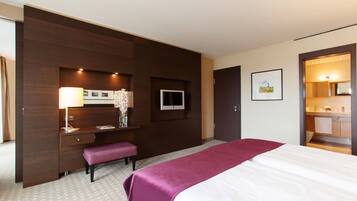 Habitación de hotel con cama, escritorio con lámpara, televisor y baño visible a través de una puerta abierta.