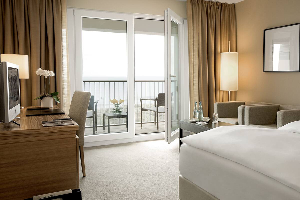 A hotel room with a bed, a desk with a computer, a chair, a small table with a vase of flowers, and a balcony with a chair.
