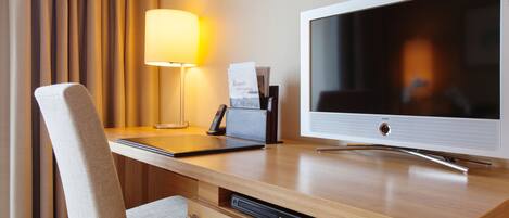Habitación de hotel con un escritorio de madera, televisión de pantalla plana, lámpara, silla y refrigerador pequeño.