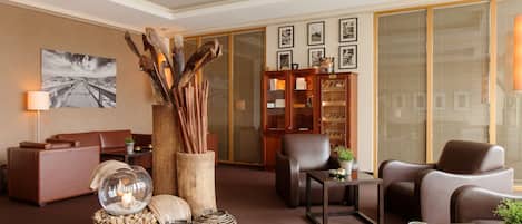 A modern living room with a sofa, armchairs, a glass coffee table, and a fireplace. There are decorative items like a glass dome, a wooden sculpture, and a bookshelf.
