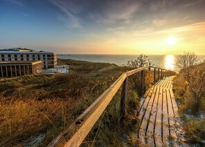 A wooden boardwalk leading to a building with a pool and a view of the ocean at sunset.
