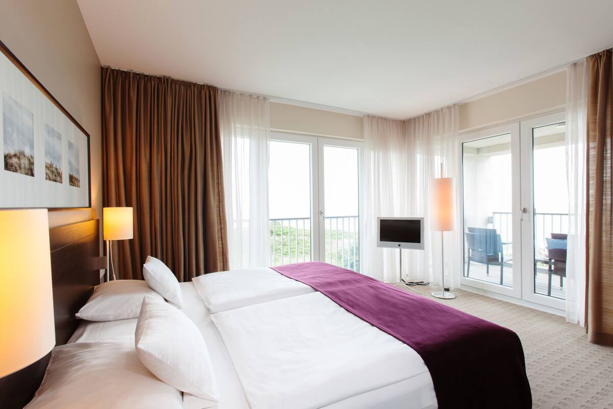 A hotel room with a large bed, a flat-screen TV, a balcony with outdoor seating, and a large window with curtains.