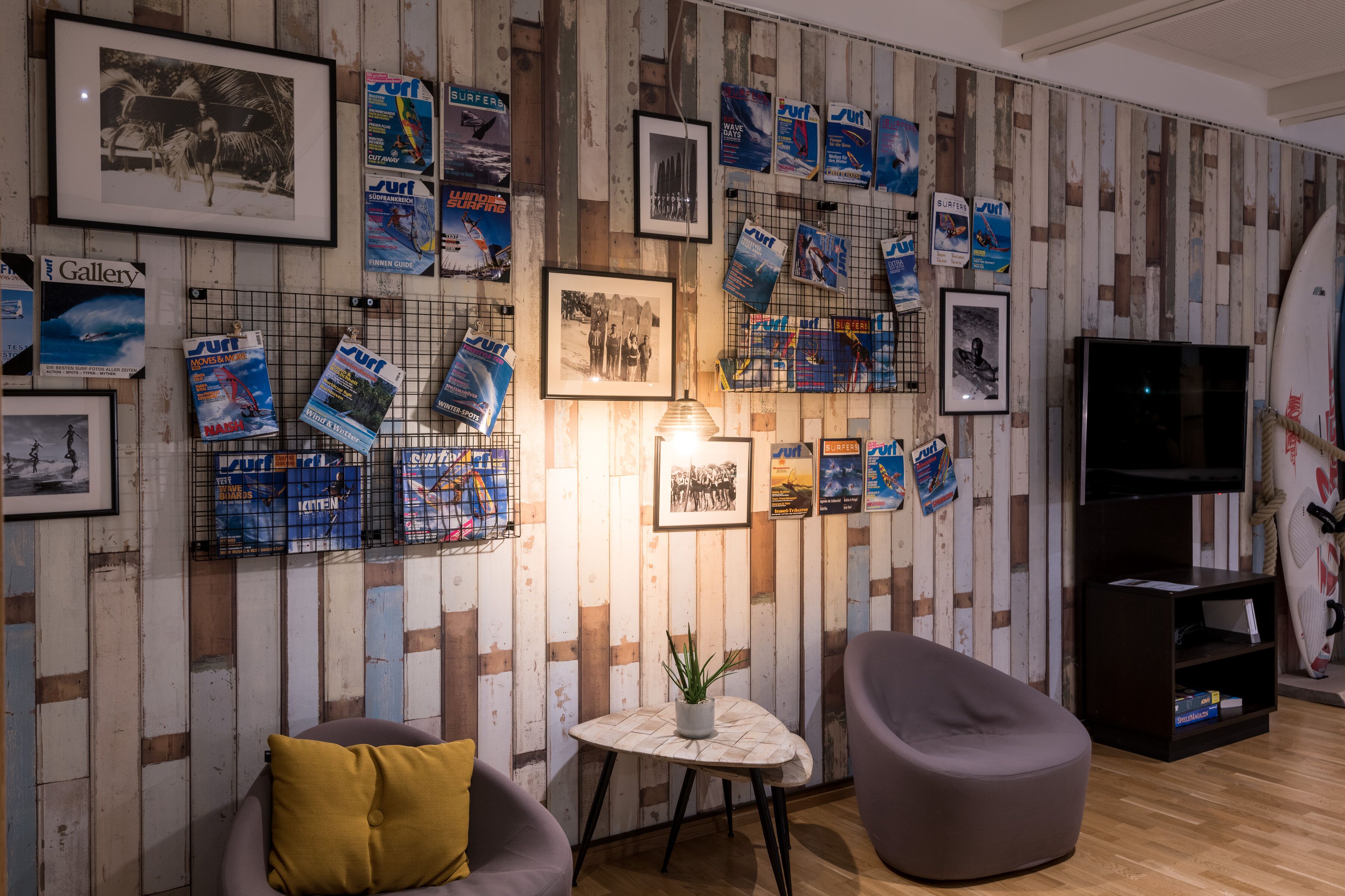 Habitación con pared de madera decorada con revistas de surf y fotografías enmarcadas, una mesita con una planta y un sofá oscuro con un cojín amarillo.