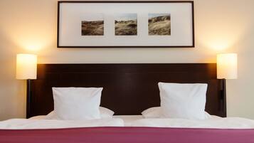 A neatly made bed with white linens and two white pillows, flanked by two wall-mounted lamps. Above the bed, there is a framed picture of a landscape.