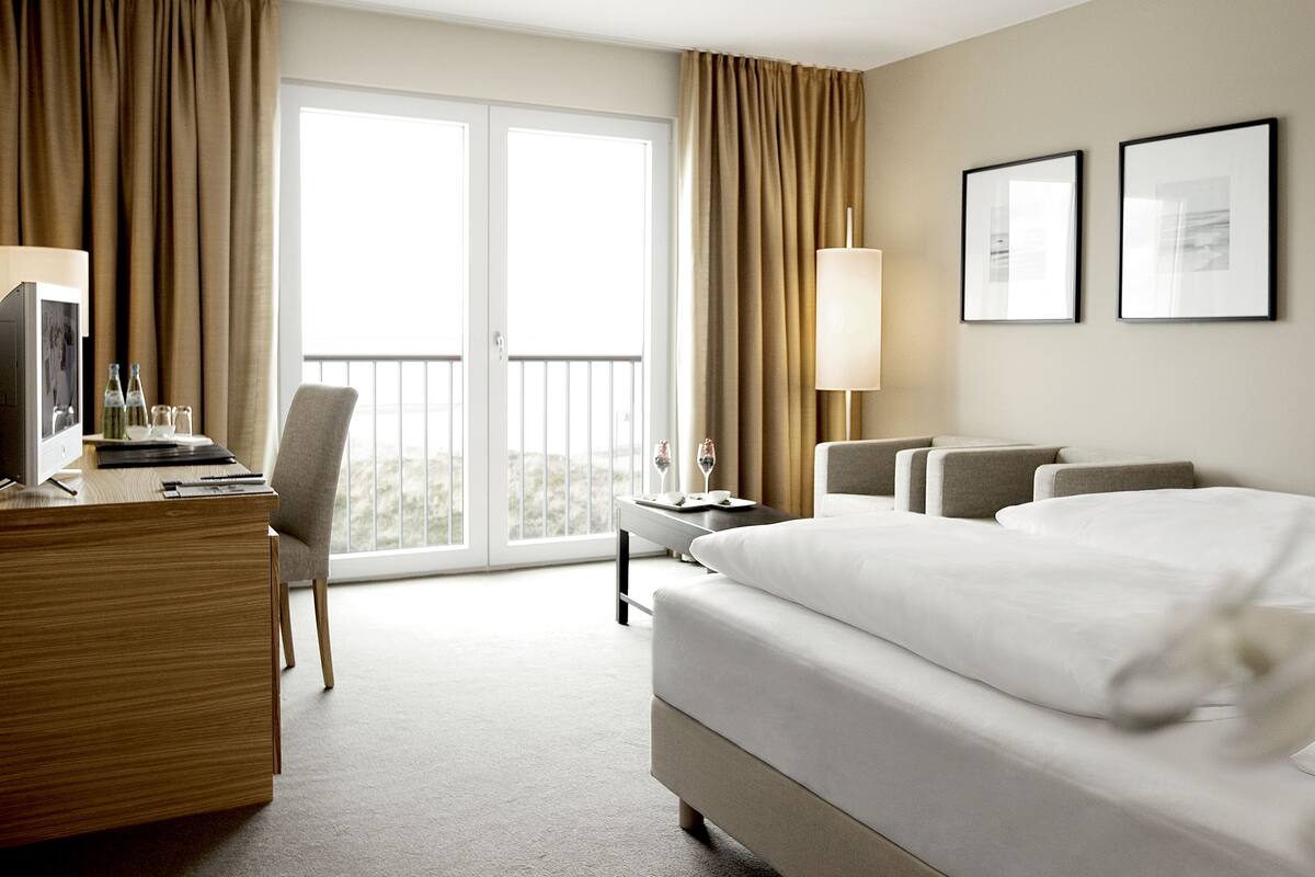 A hotel room with a bed, desk, chair, and a balcony with curtains.