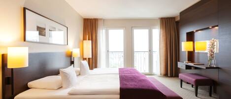 Ein Hotelzimmer mit einem großen Bett, zwei Nachttischlampen, einer Bank, einem Schreibtisch mit Lampe und einem großen Fenster mit Vorhängen.