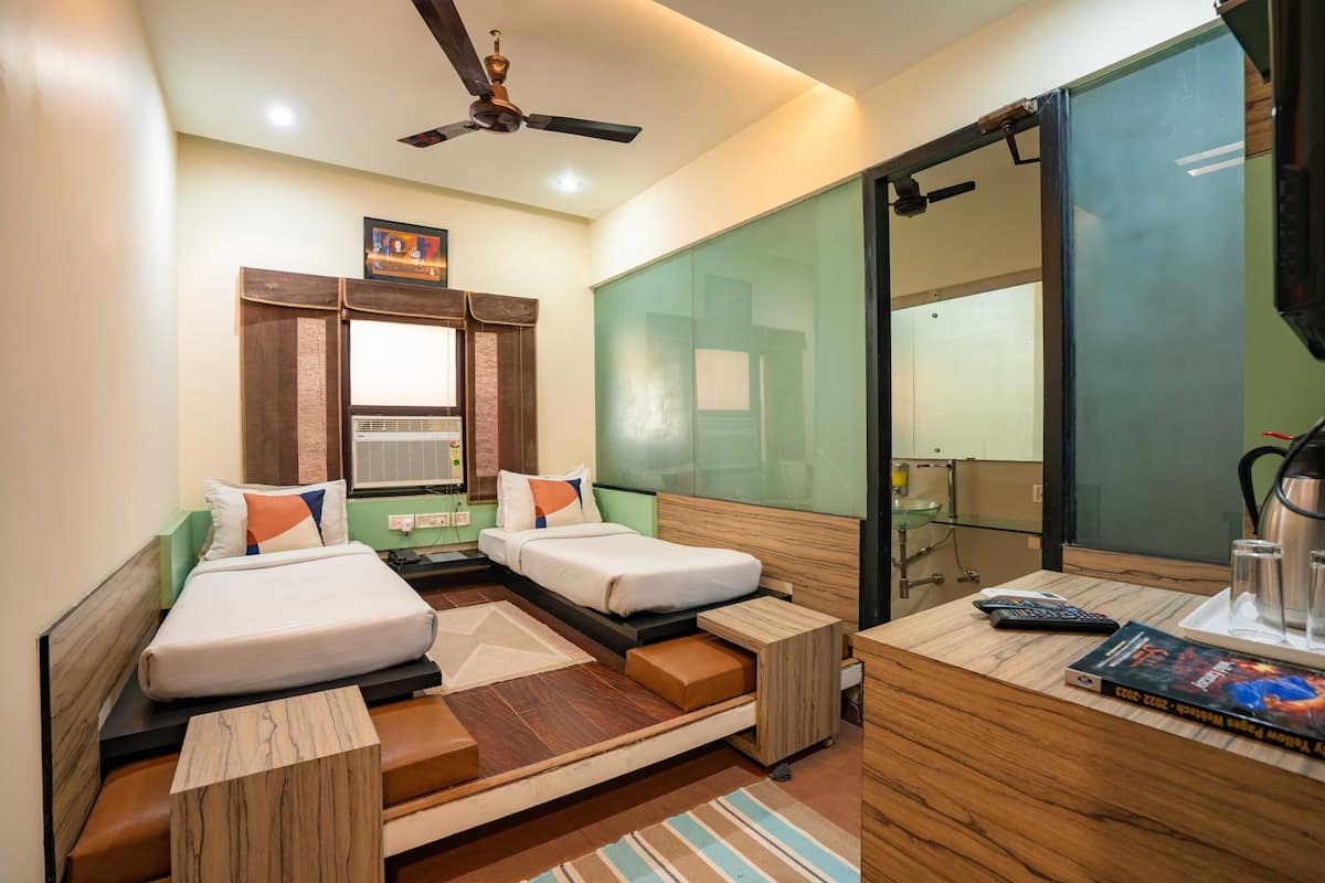 A compact hotel room with two single beds, a small desk with a chair, a mirror, a ceiling fan, and a window with blinds.
