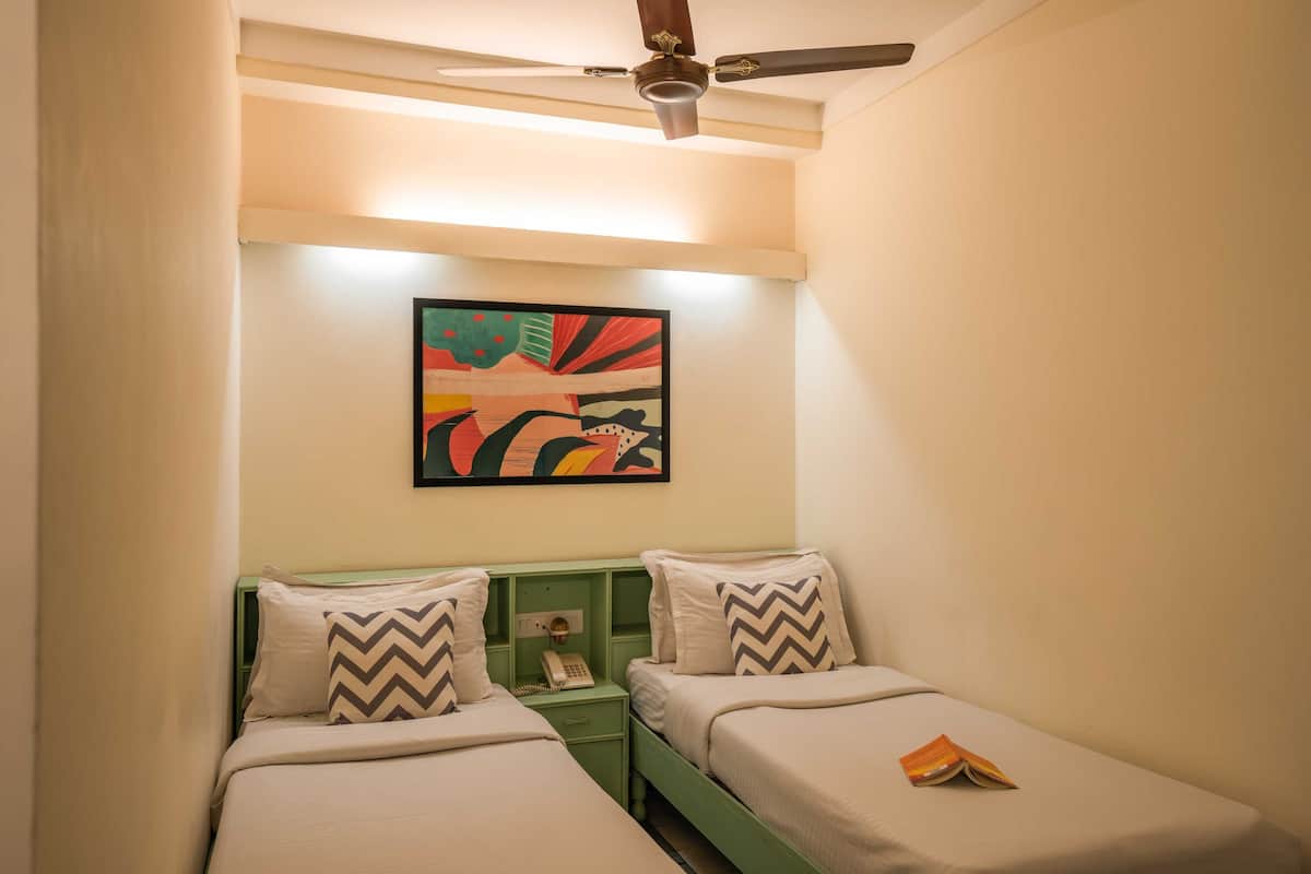 A small, well-lit room with two single beds, a green nightstand, a ceiling fan, and a colorful abstract painting on the wall.