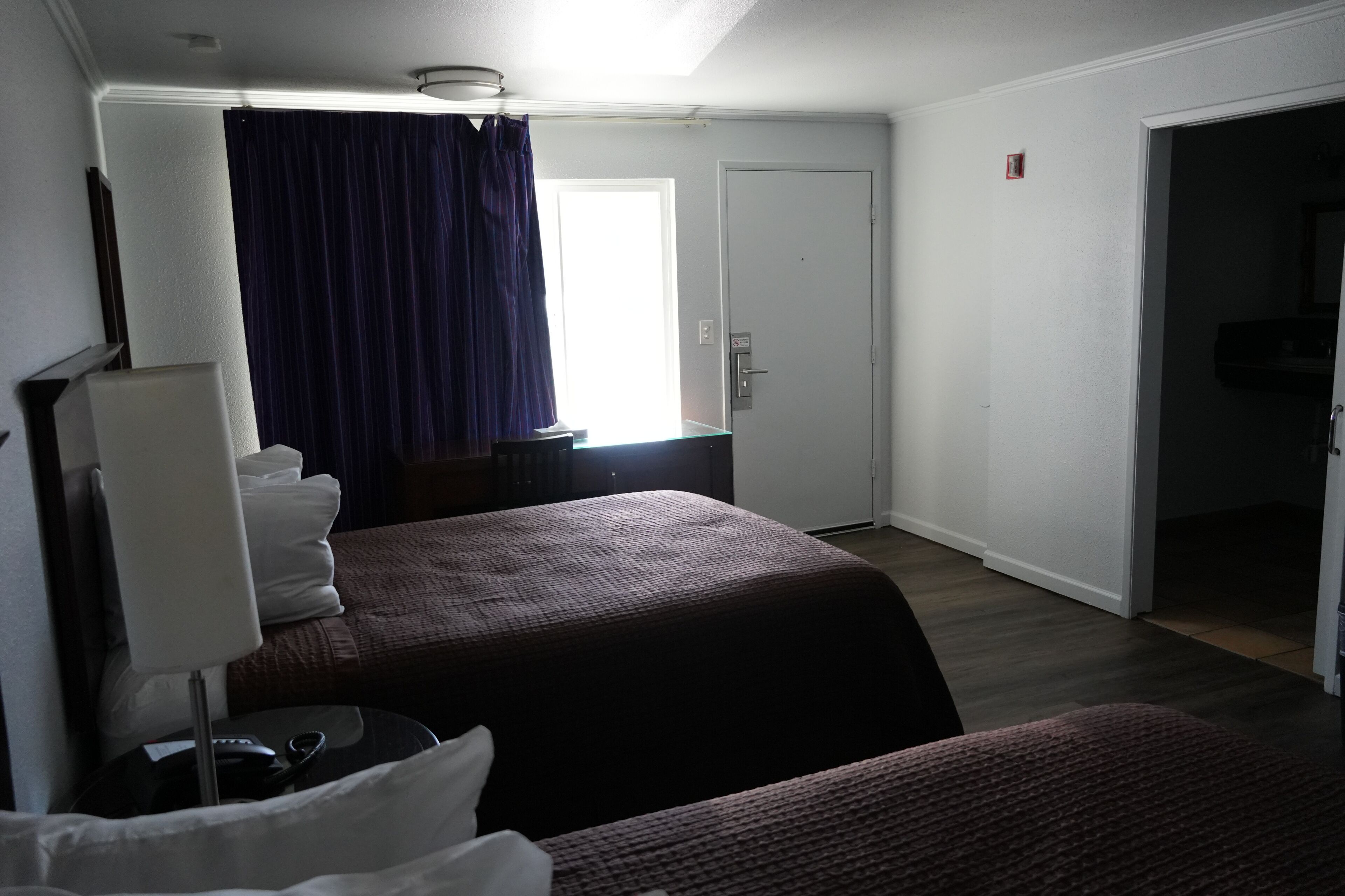 Habitación de hotel con cama, escritorio, silla, lámpara, ventana con cortinas y puerta.