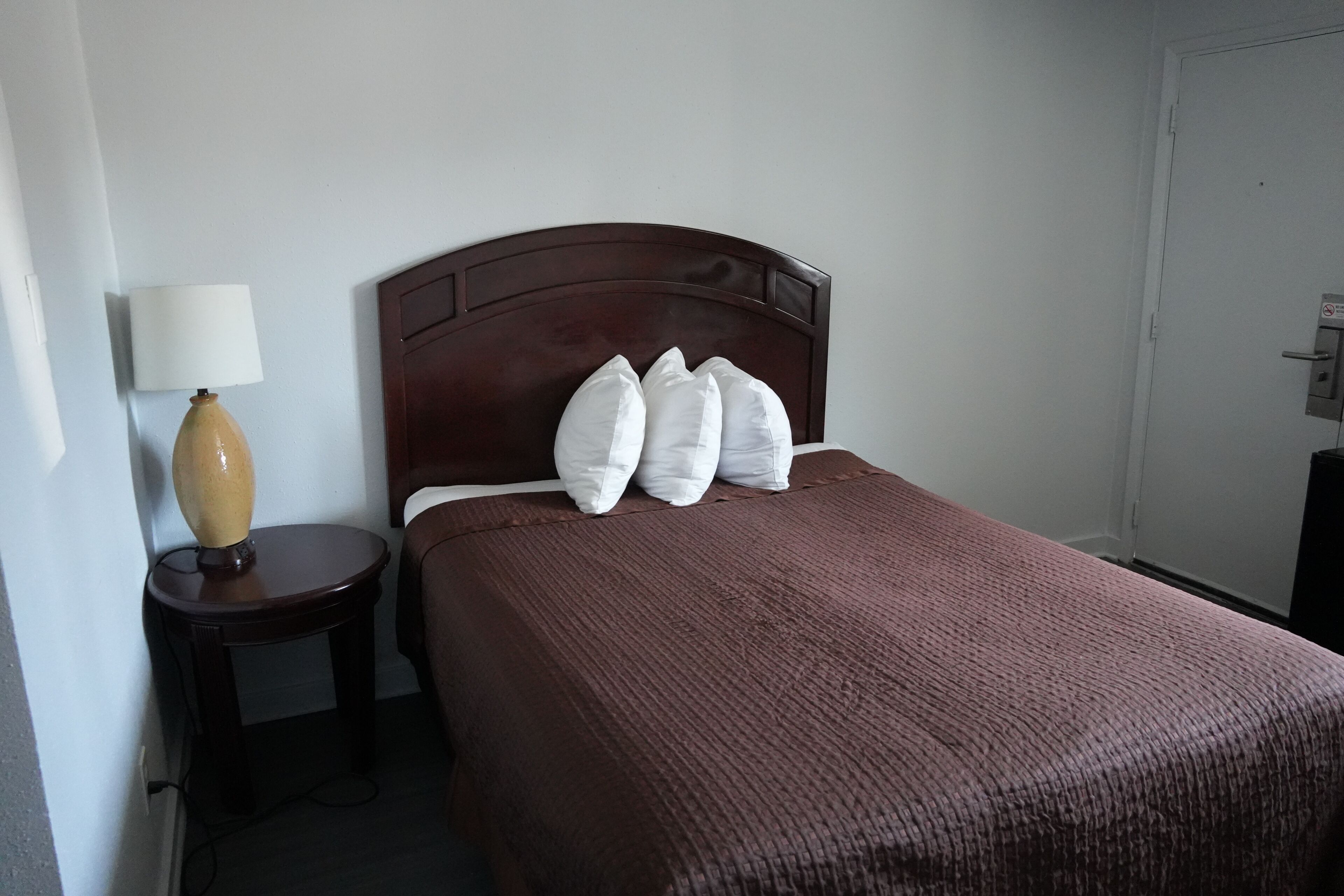 A neatly made bed with a brown bedspread and white pillows, a small wooden nightstand with a lamp, and a door to the right.