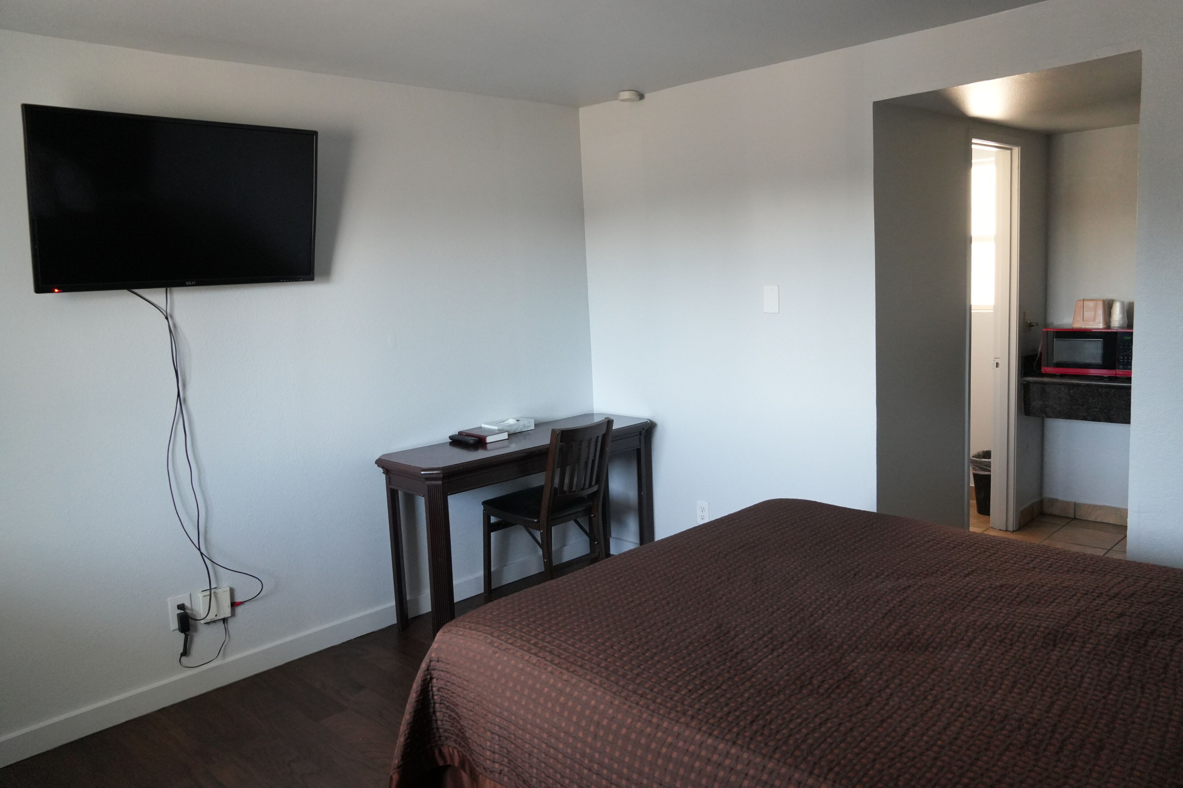 A bedroom with a bed, a desk with a chair, a television mounted on the wall, and a microwave in the adjacent room.