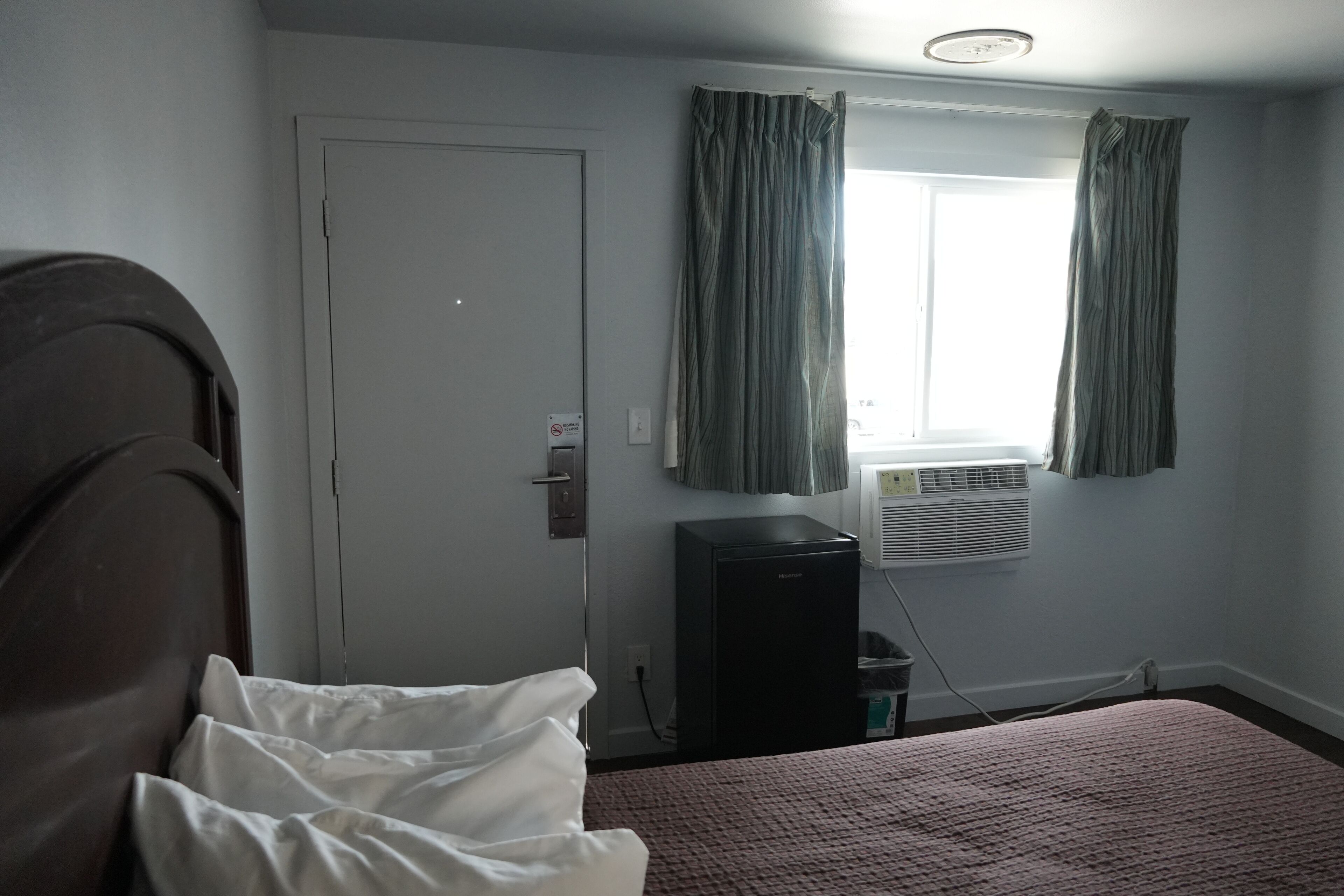 Una habitación con cama, puerta, ventana con cortinas, un equipo de aire acondicionado y un electrodoméstico pequeño y negro.