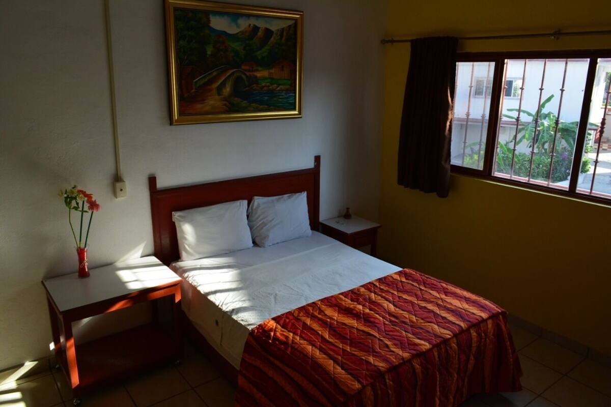 Een slaapkamer met een bed, een nachtkastje, een schilderij aan de muur en een raam met gordijnen.