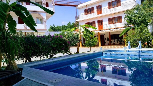 A swimming pool with a clear blue water surface, surrounded by lush greenery and a white building with balconies. - Hotel Hacienda de Zapata (Jiutepec)