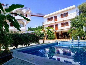 Una piscina con superficie de agua azul cristalina, rodeada de abundante follaje y un edificio blanco con balcones.