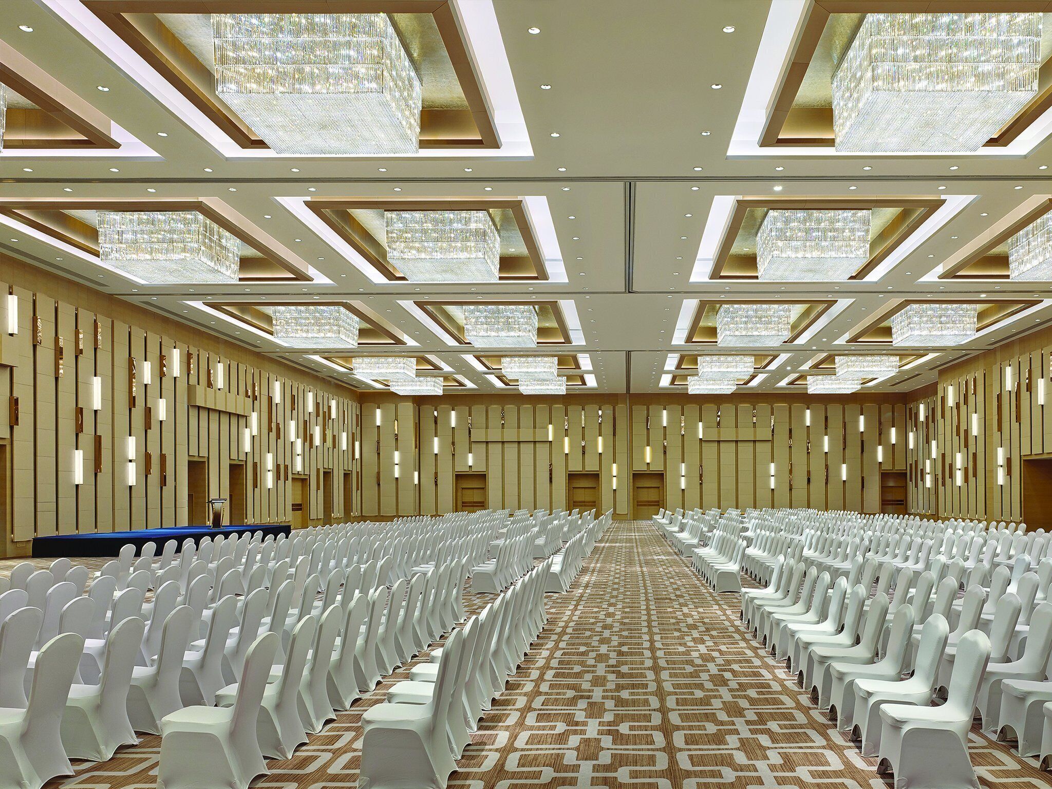 A large conference room with rows of white chairs, a patterned carpet, and a modern chandelier ceiling.