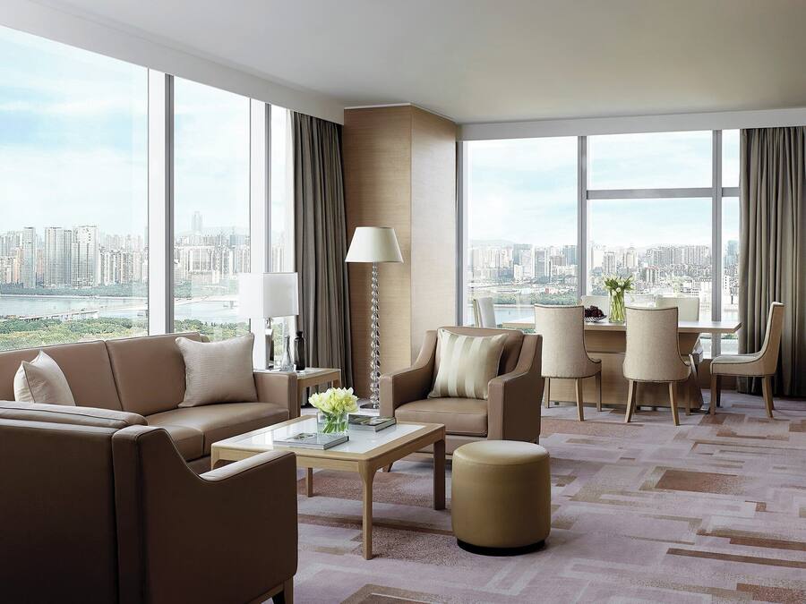 A modern hotel room with a city view, a sofa, a coffee table, and a dining area.