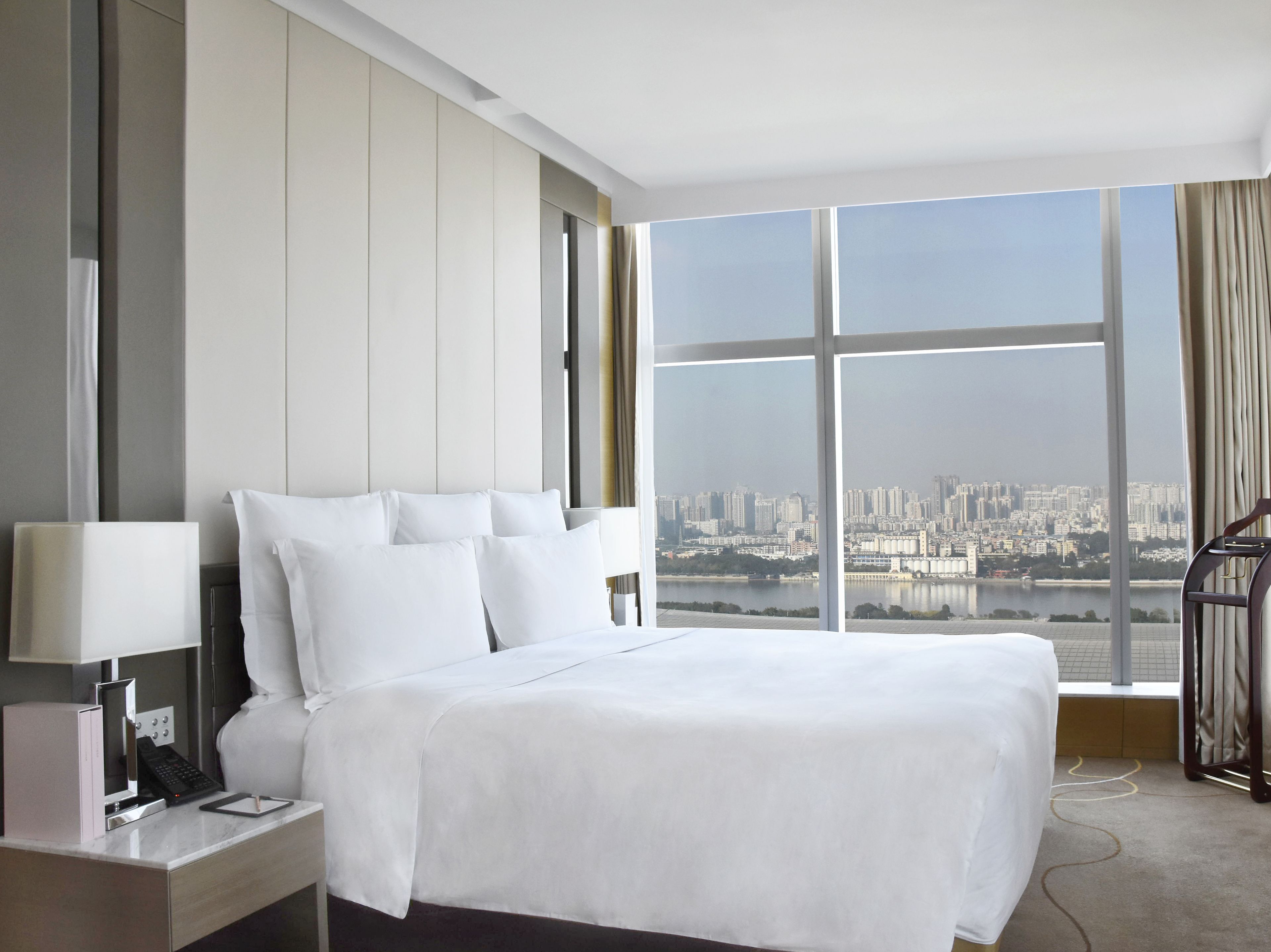 Une chambre d’hôtel avec un grand lit, une table de chevet avec une lampe, un téléphone et une vue sur le paysage urbain grâce à de grandes fenêtres.