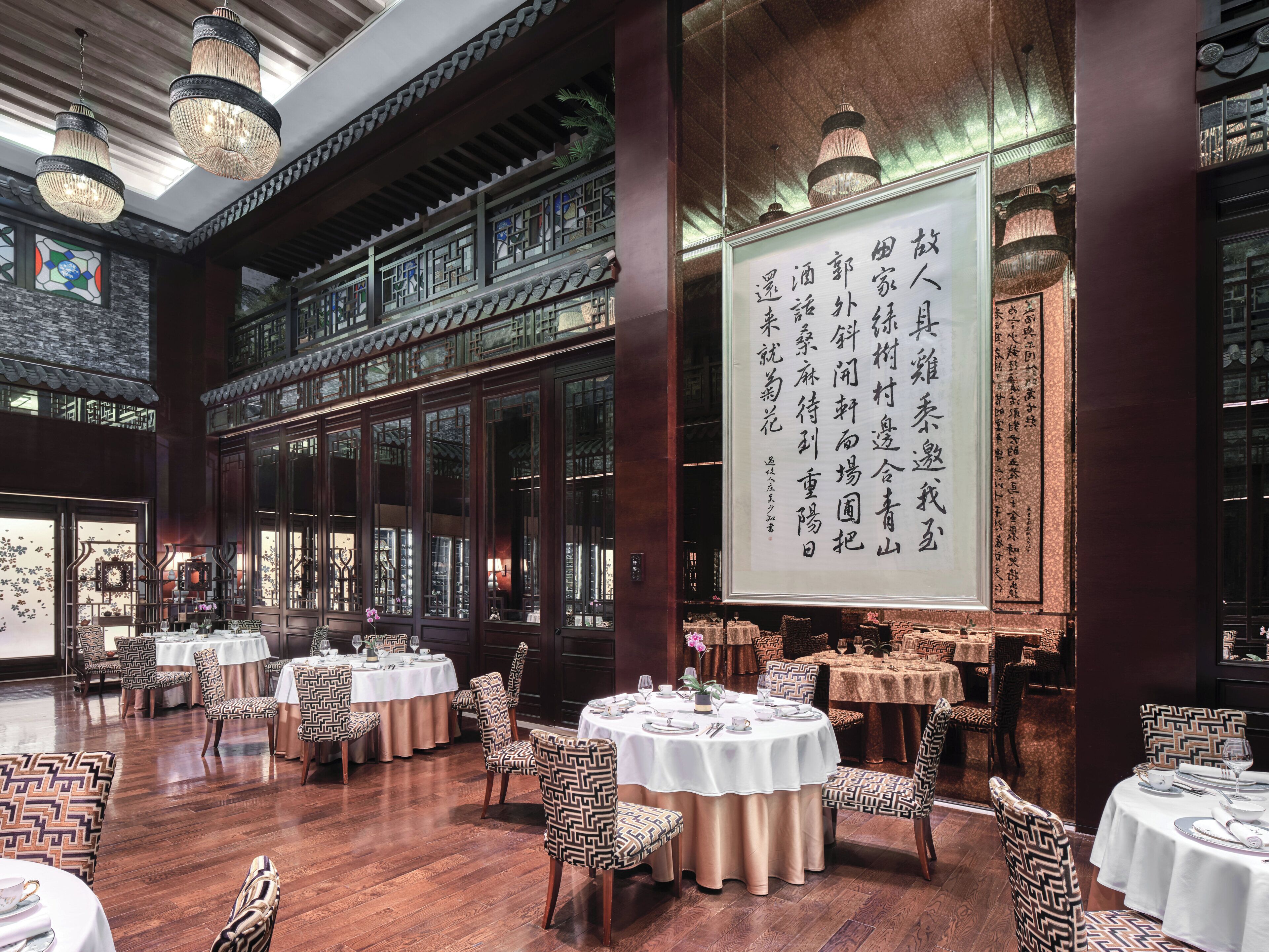 Uma sala de jantar tradicional com mesas redondas, cadeiras estampadas e um grande painel vertical com texto.