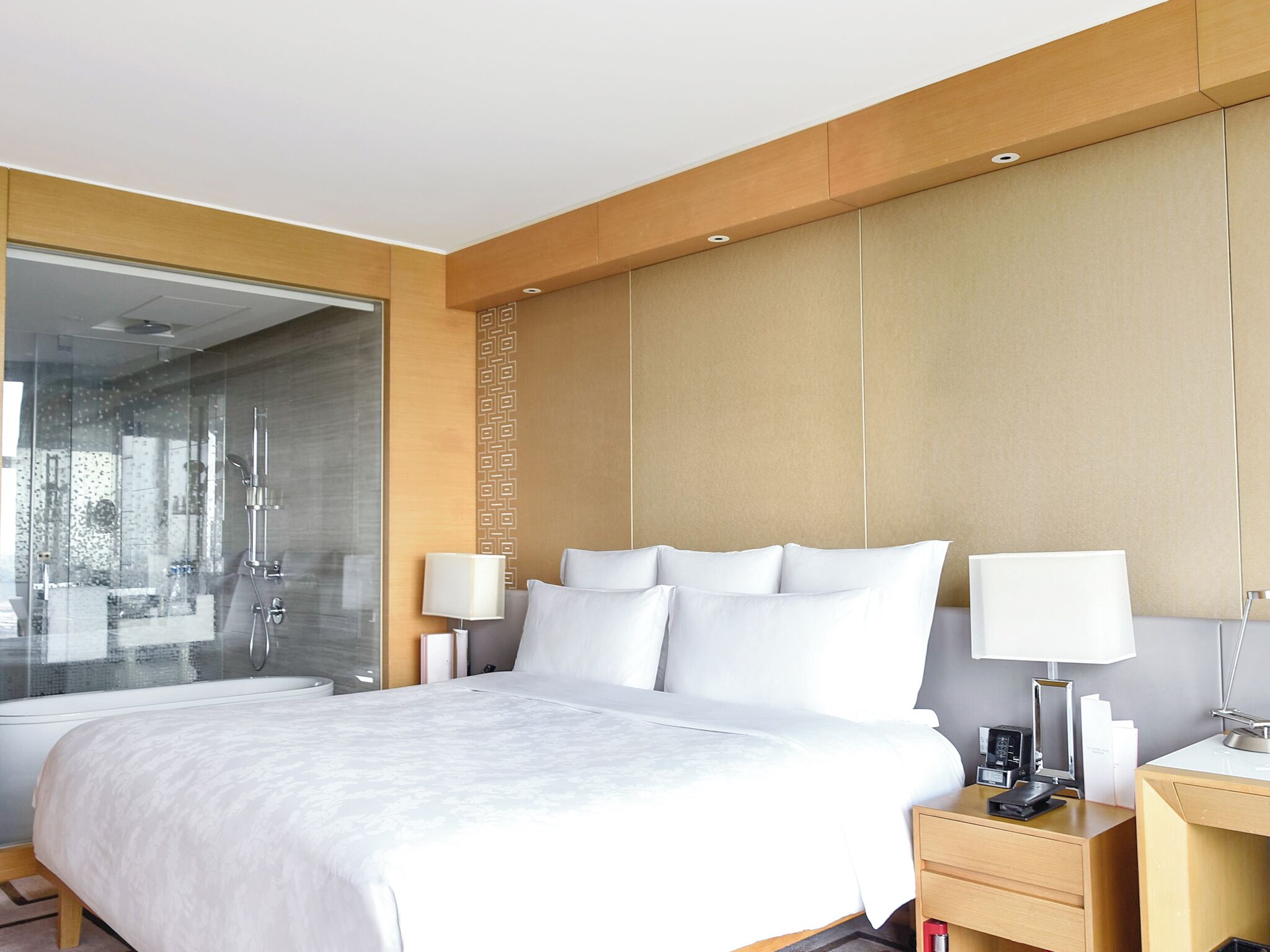 A modern hotel room with a large bed, bedside tables, a glass shower enclosure, and a wooden headboard.