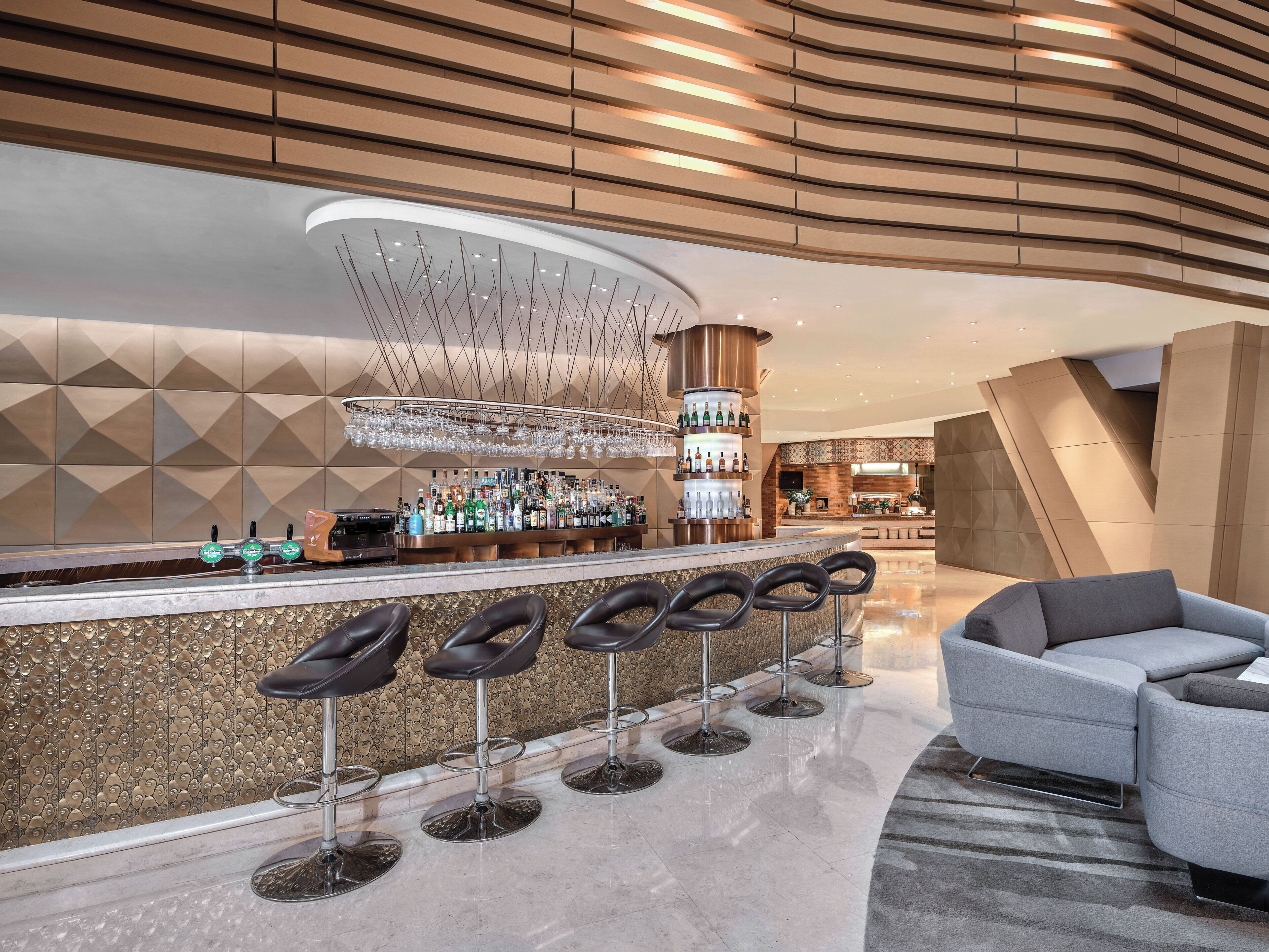 Zona de bar moderna con barra curva, varios taburetes y una gran variedad de botellas y copas.