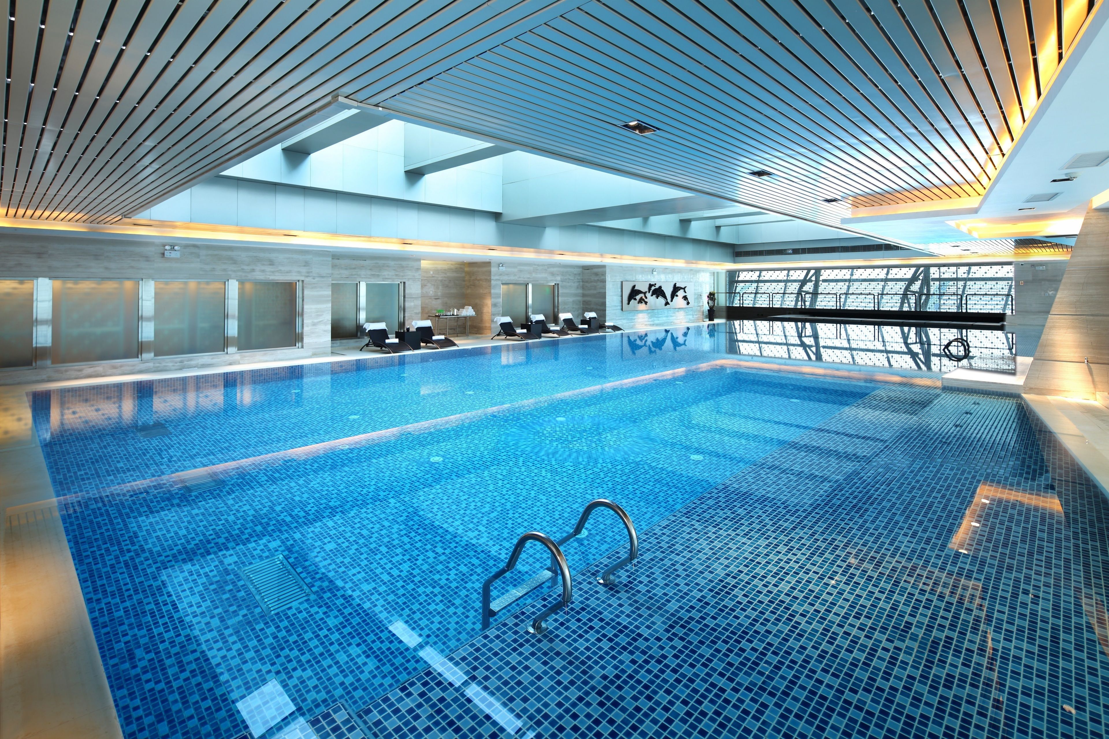 A large indoor swimming pool with a tiled floor, a ceiling with horizontal beams, and a glass wall on one side.