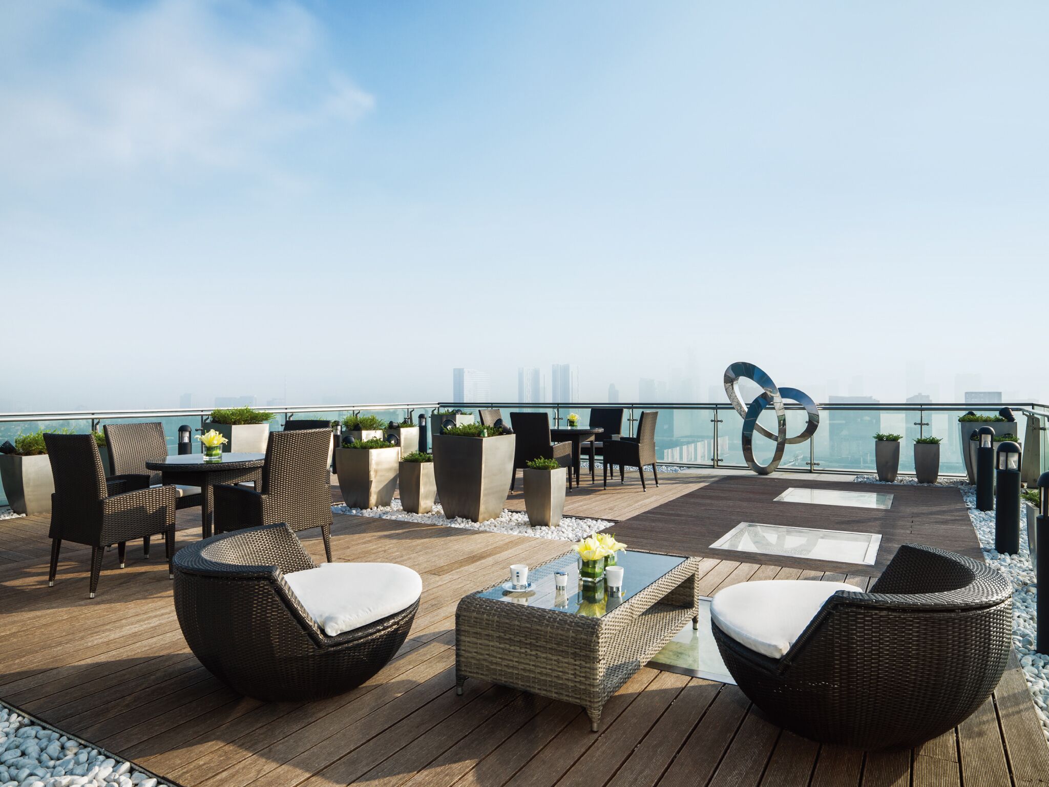 A rooftop terrace with wicker furniture, potted plants, and a glass balustrade overlooking a cityscape.