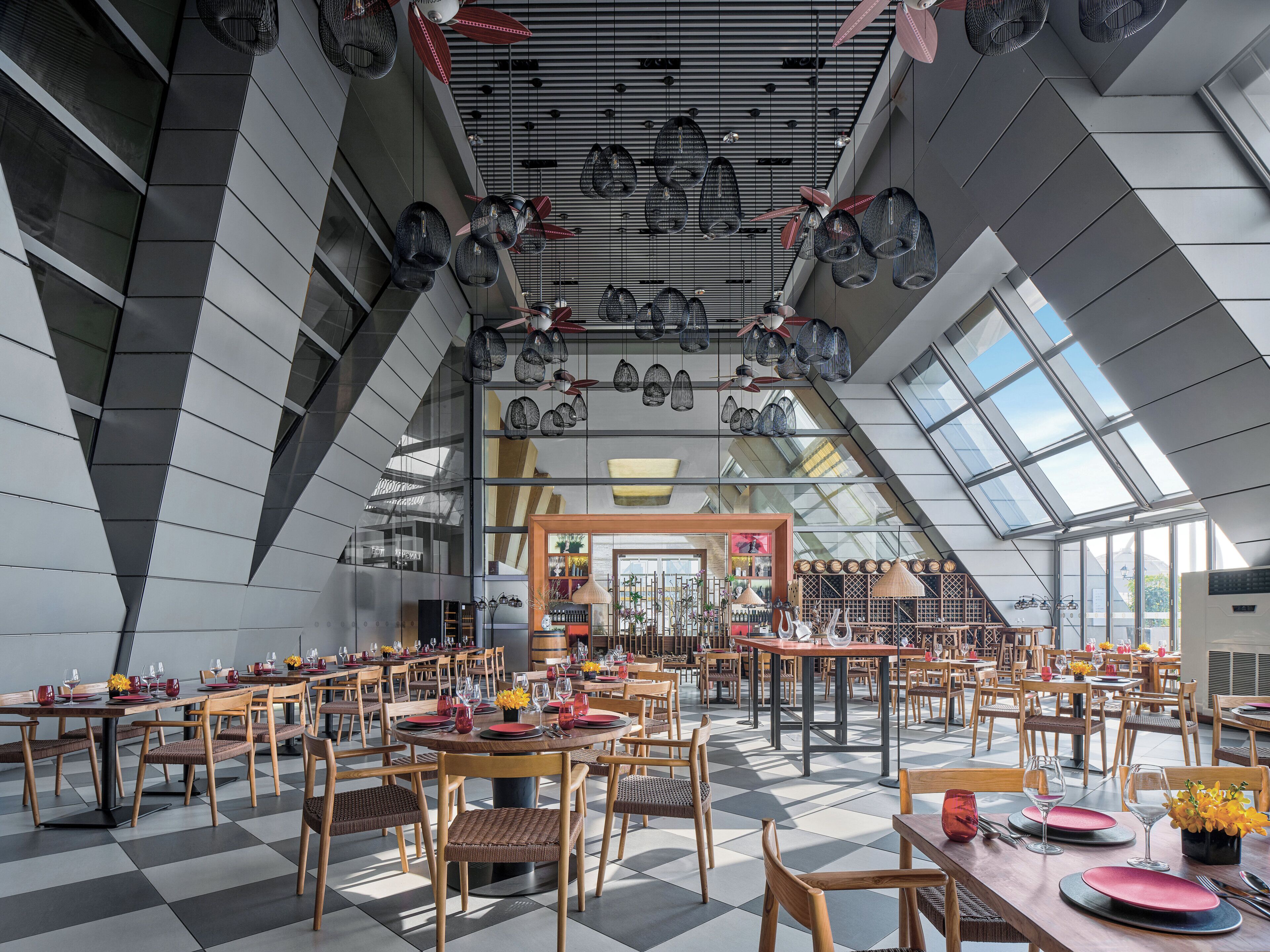 Un vaste espace dédié à la salle à manger, avec de nombreuses tables et chaises, un plafond haut agrémenté de luminaires suspendus et de grandes fenêtres laissant entrer la lumière naturelle.