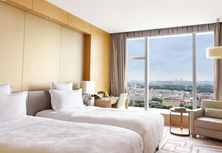 A hotel room with two beds, a desk, a chair, and a large window offering a city view.