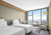 A hotel room with two beds, a desk, a chair, and a view of the city.