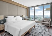 A hotel room with a large bed, a desk, a chair, and a view of a cityscape through large windows.