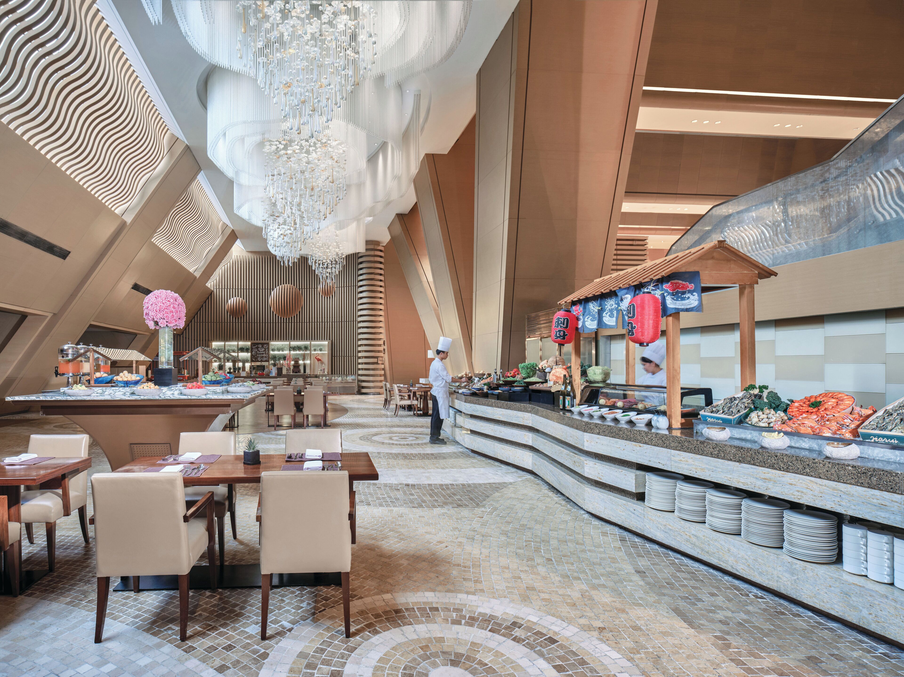 A modern hotel lobby with a central seating area, a food display, and a high ceiling with a decorative chandelier.