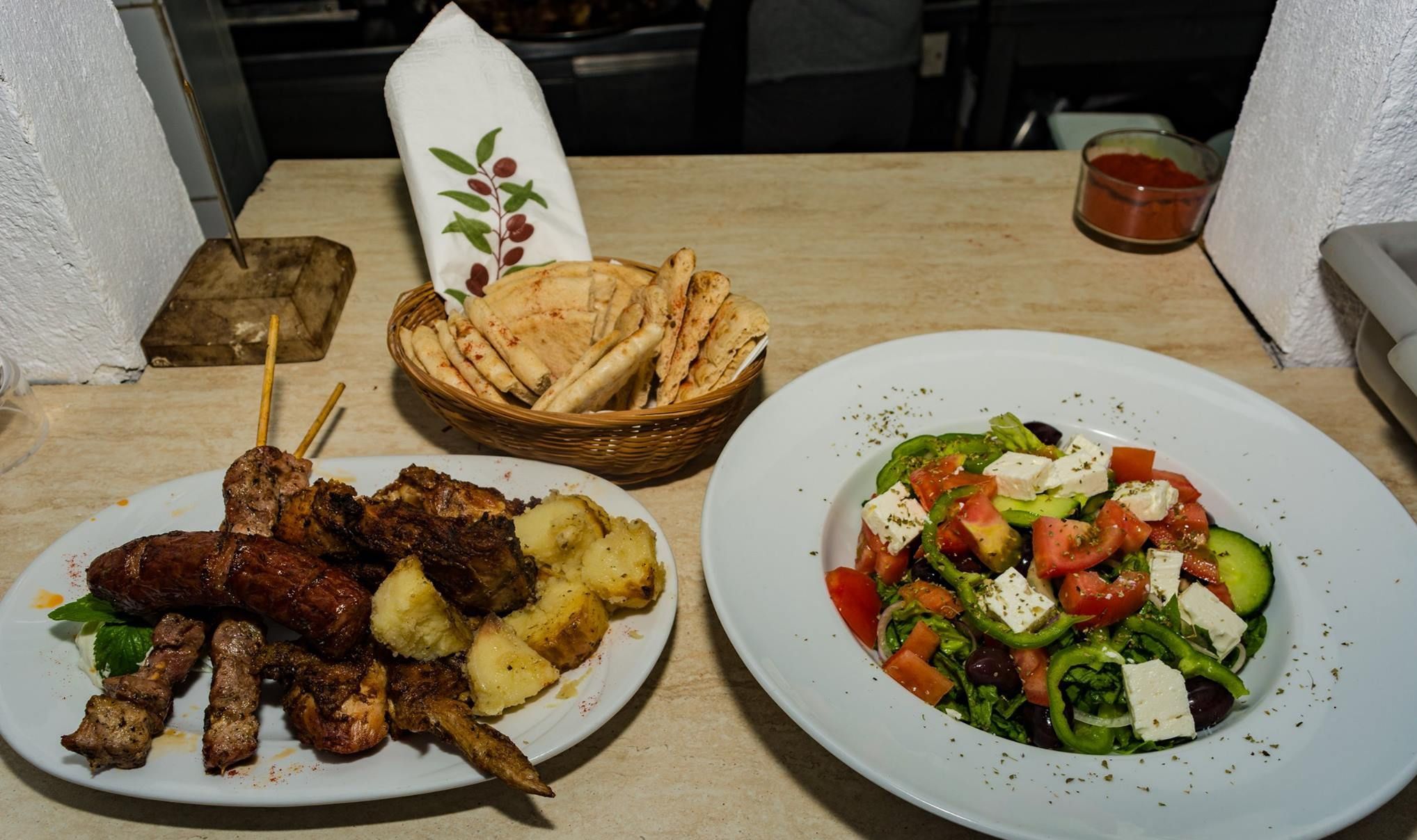 Una mesa con platos de carne a la parrilla, patatas asadas y una ensalada griega.