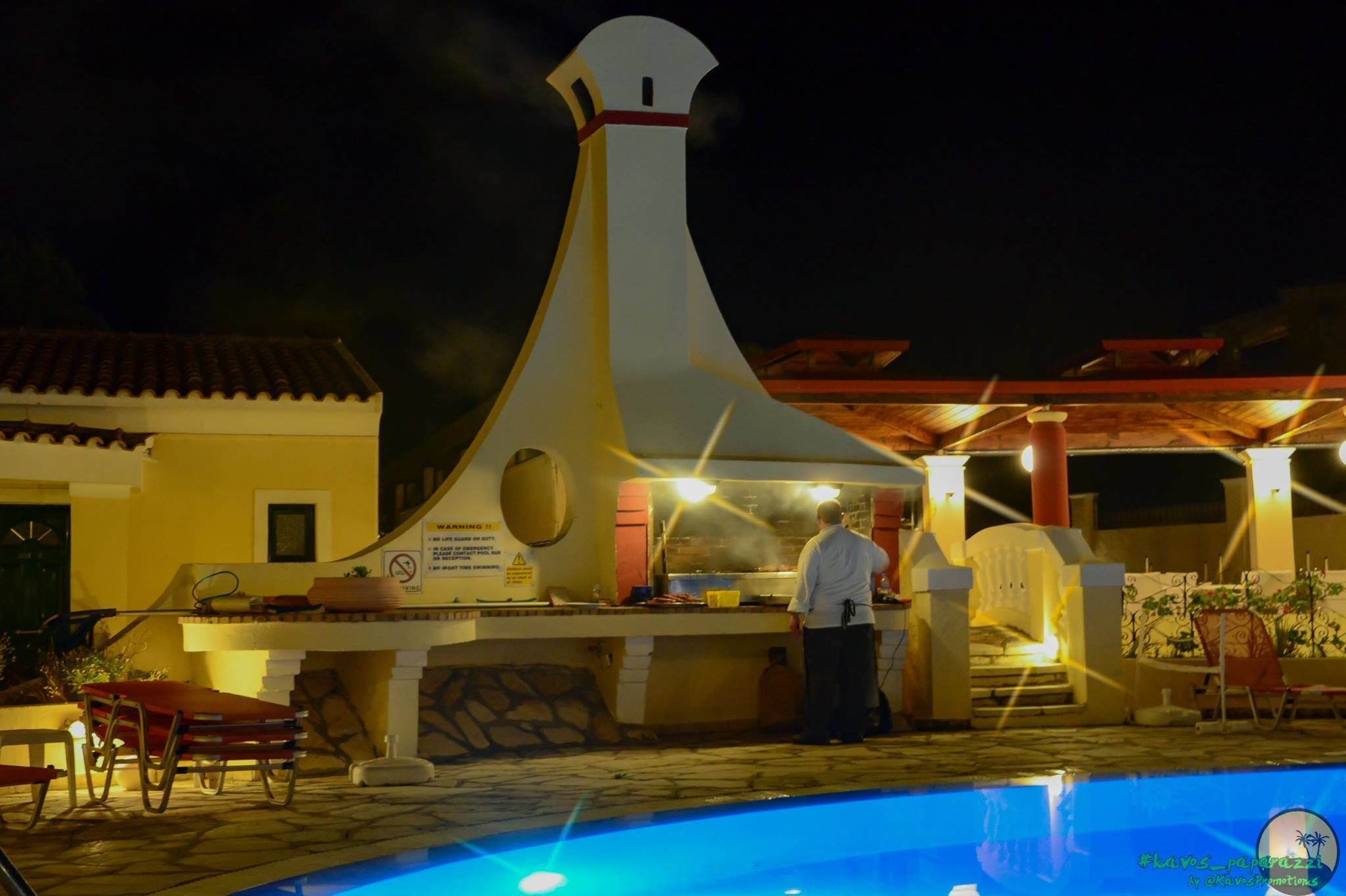 An outdoor bar area with a chef preparing food, a pool, and a dining setup with chairs and tables.