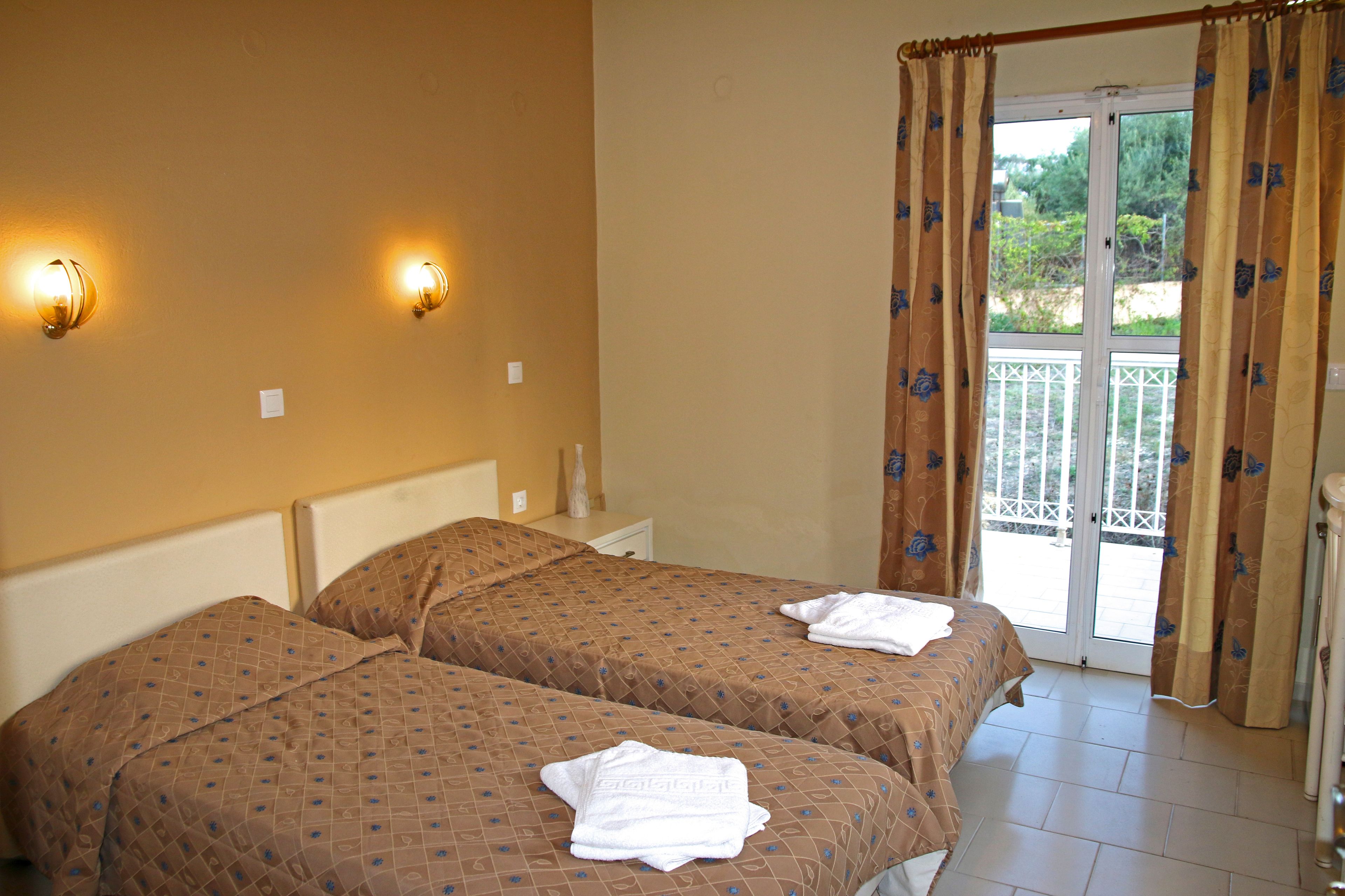 Quarto de hotel com duas camas, varanda com guarda-corrimão branco e cortinas com padrão floral.
