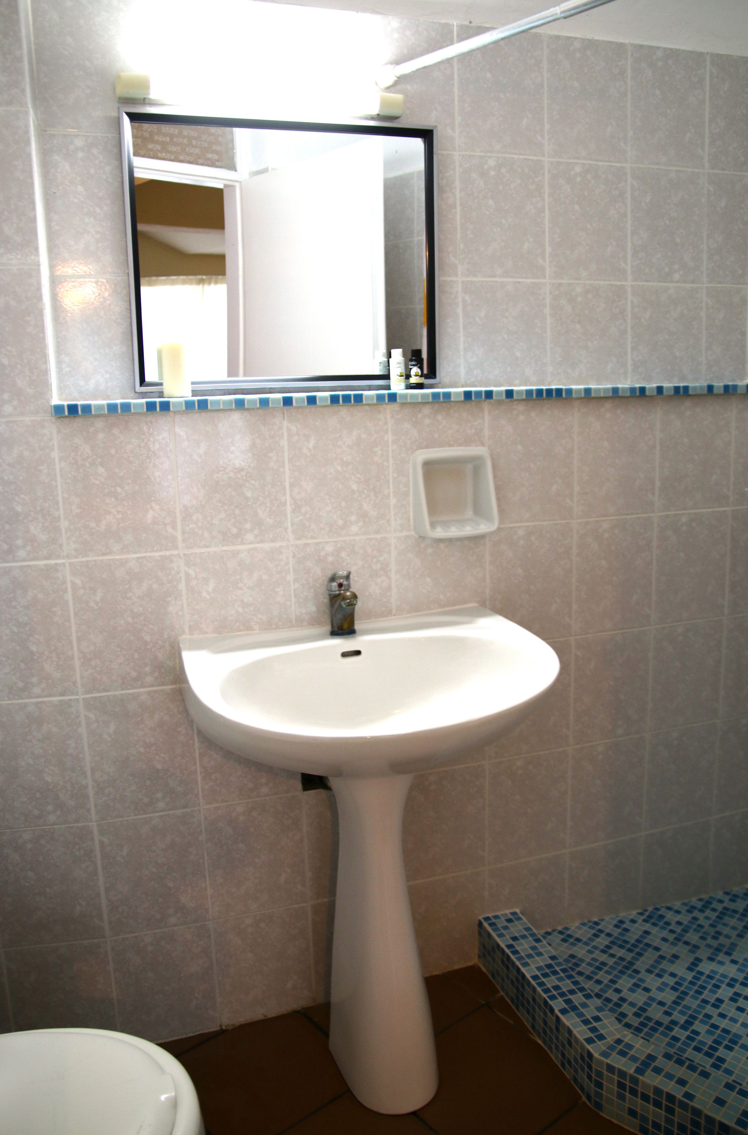 Un baño con un lavamanos pedestal blanco, un espejo encima y suelo de baldosas.
