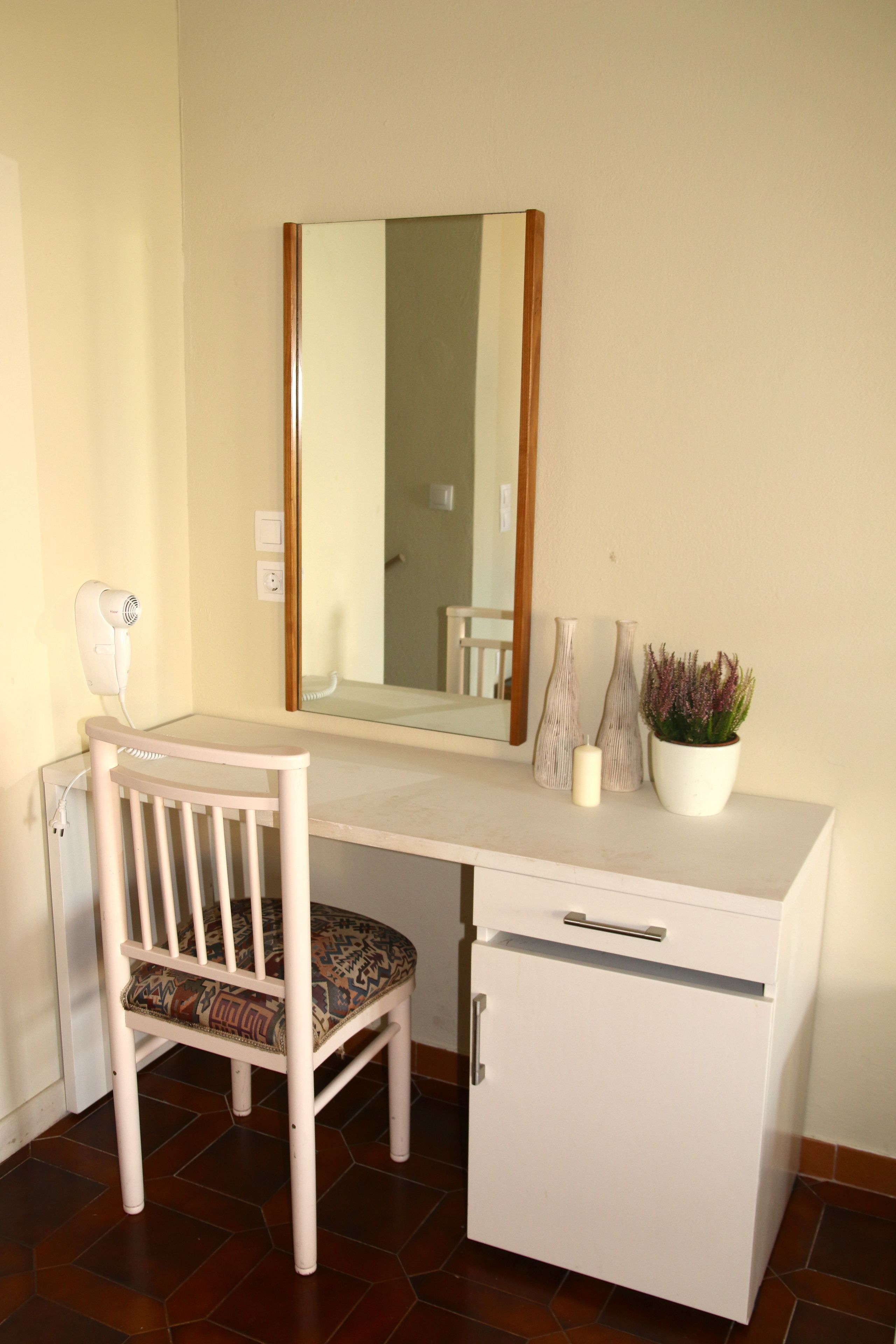 Una habitación pequeña y privada con un escritorio blanco, una silla, un espejo, una planta en maceta y un mueble bajo.