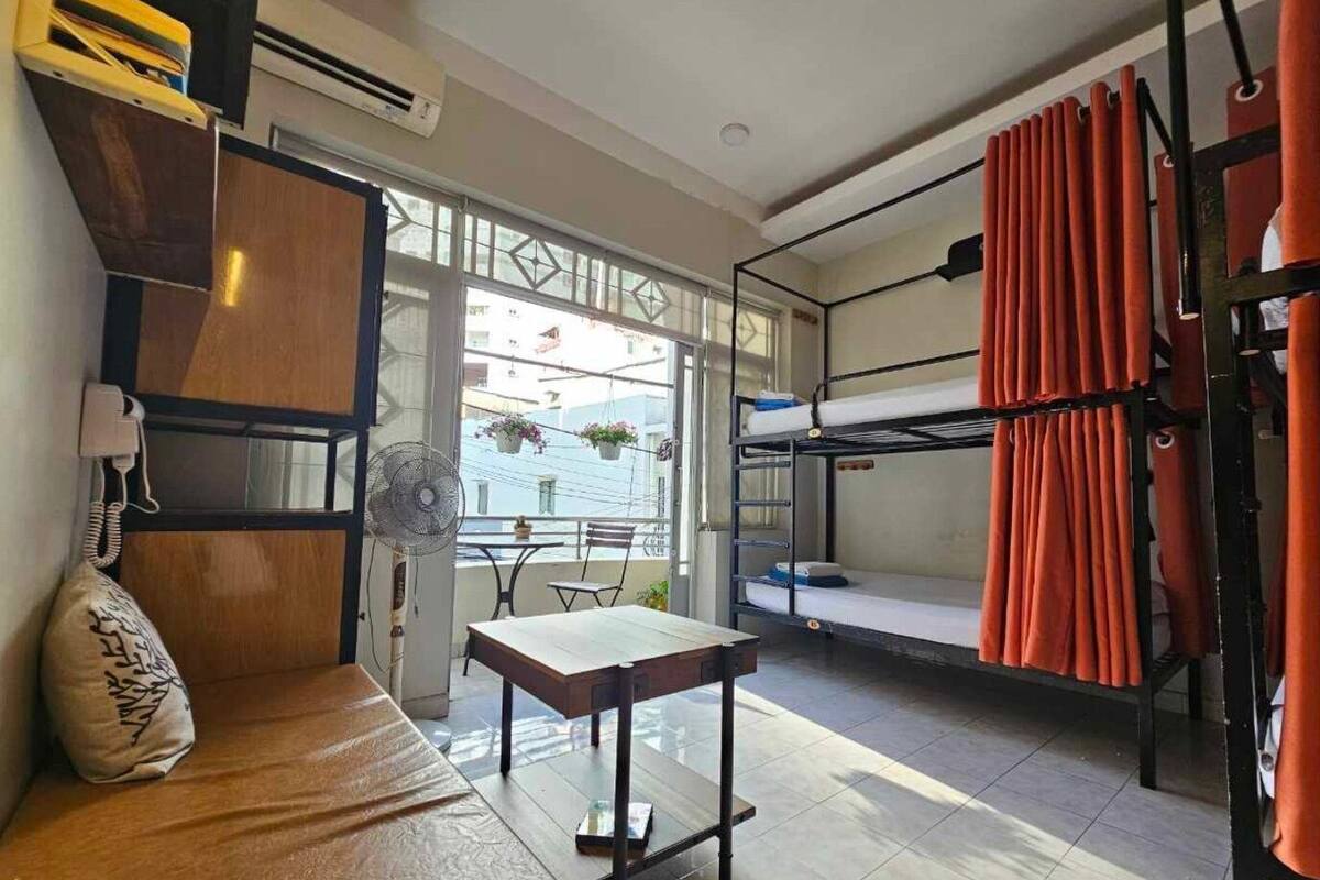 Een compacte kamer met stapelbedden, een klein tafeltje, een ventilator en een raam met uitzicht op een balkon met planten.