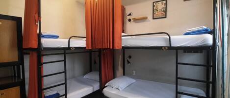 A dormitory room with bunk beds, orange curtains, a small shelf with a framed picture, and a wall-mounted lamp.