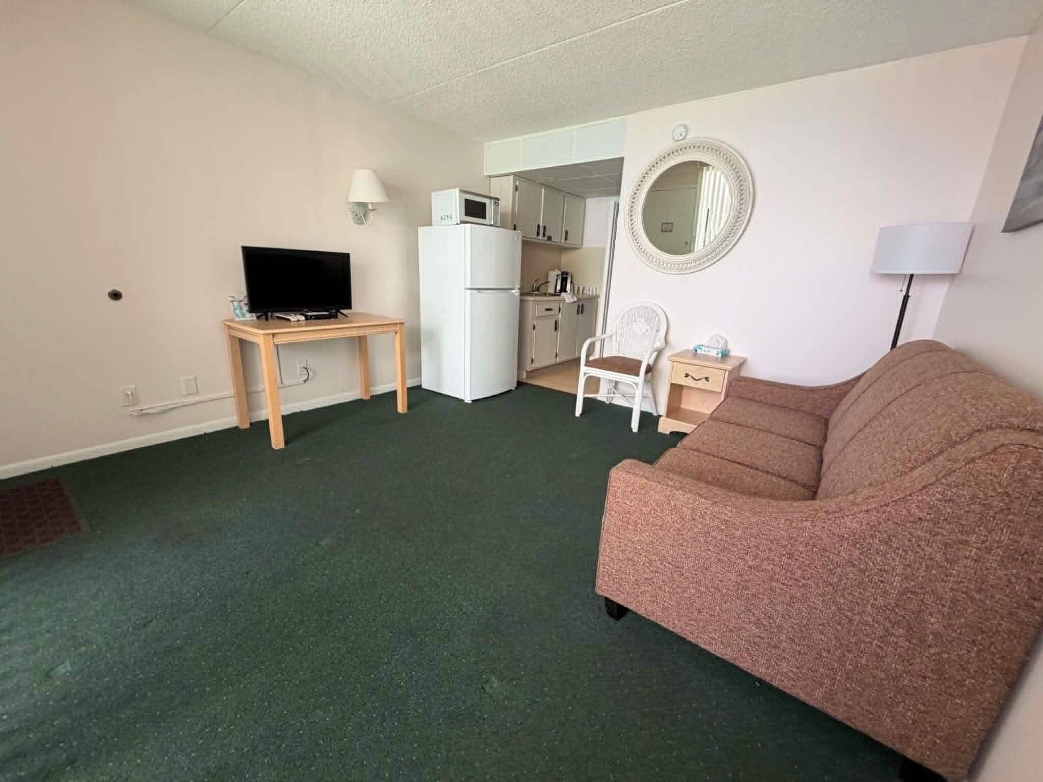 A compact living space with a sofa, a television on a small table, a kitchenette, and a mirror on the wall.